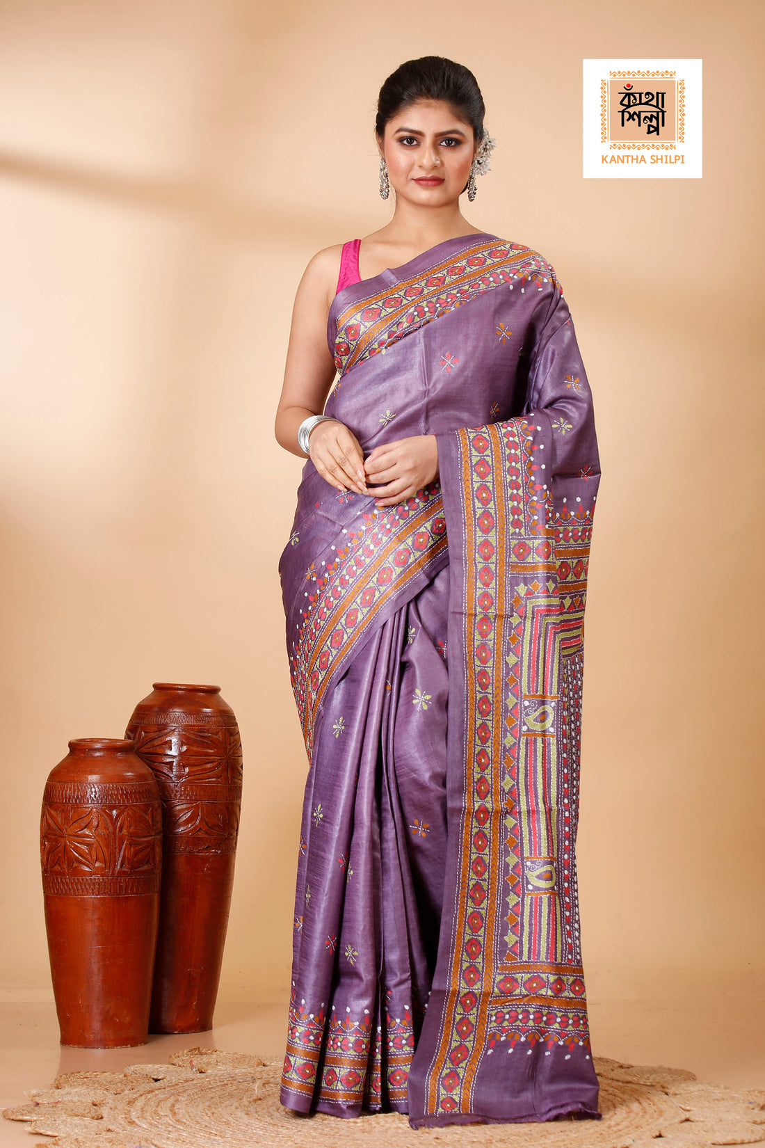 Woman wearing a purple saree with a traditional design, standing next to two brown vases on a beige background.